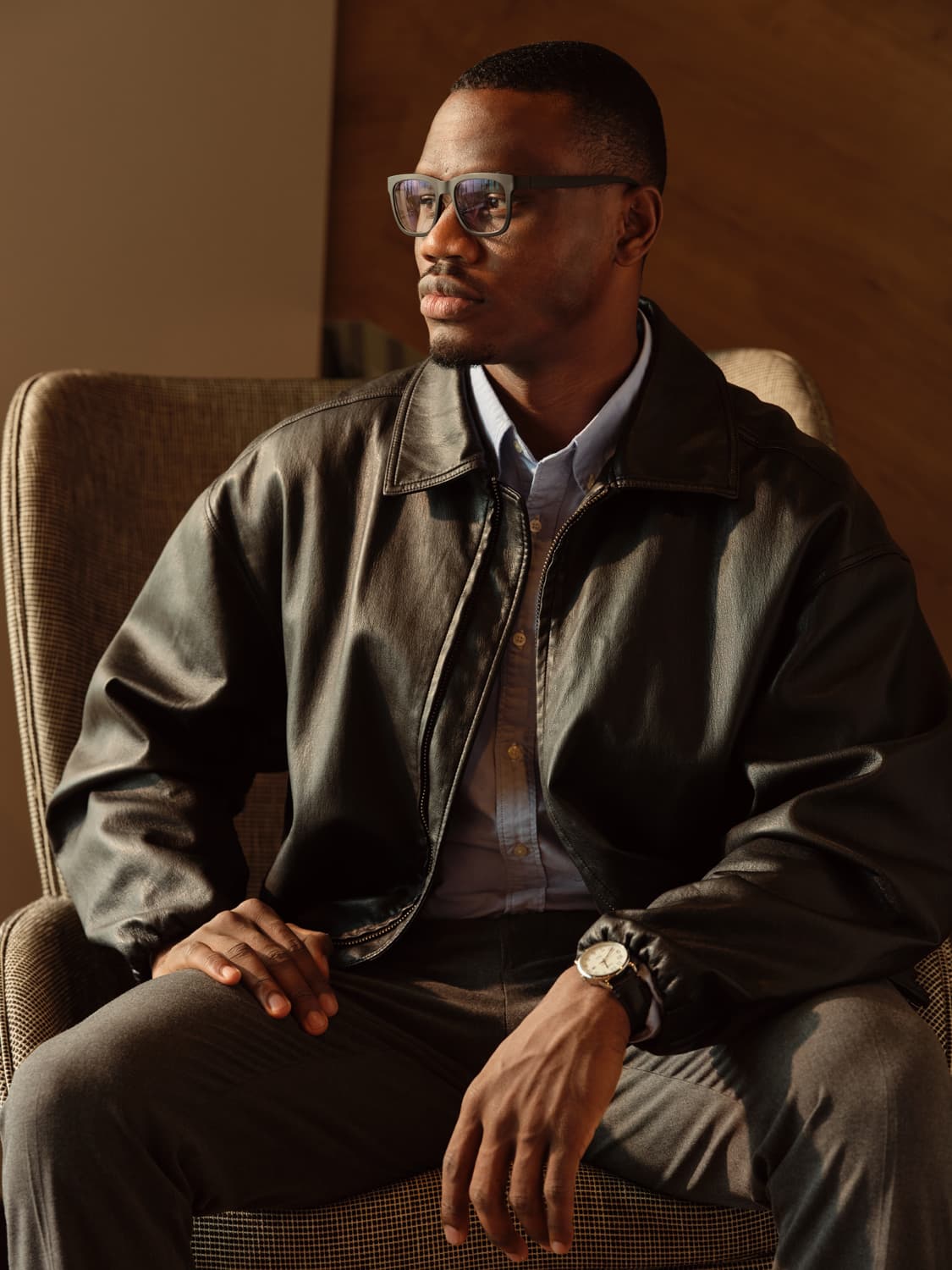 Man in clear framed glasses and a black leather bomber jacket over a light blue button down, seated in a patterned upholstered chair with hands clasped, looking off camera in warm directional light