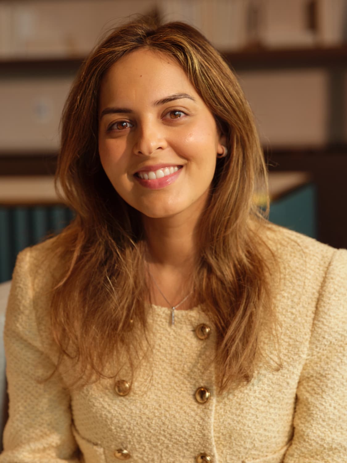 Woman with long honey brown hair in a cream tweed jacket with gold buttons, smiling warmly with soft natural light against a blurred library backdrop