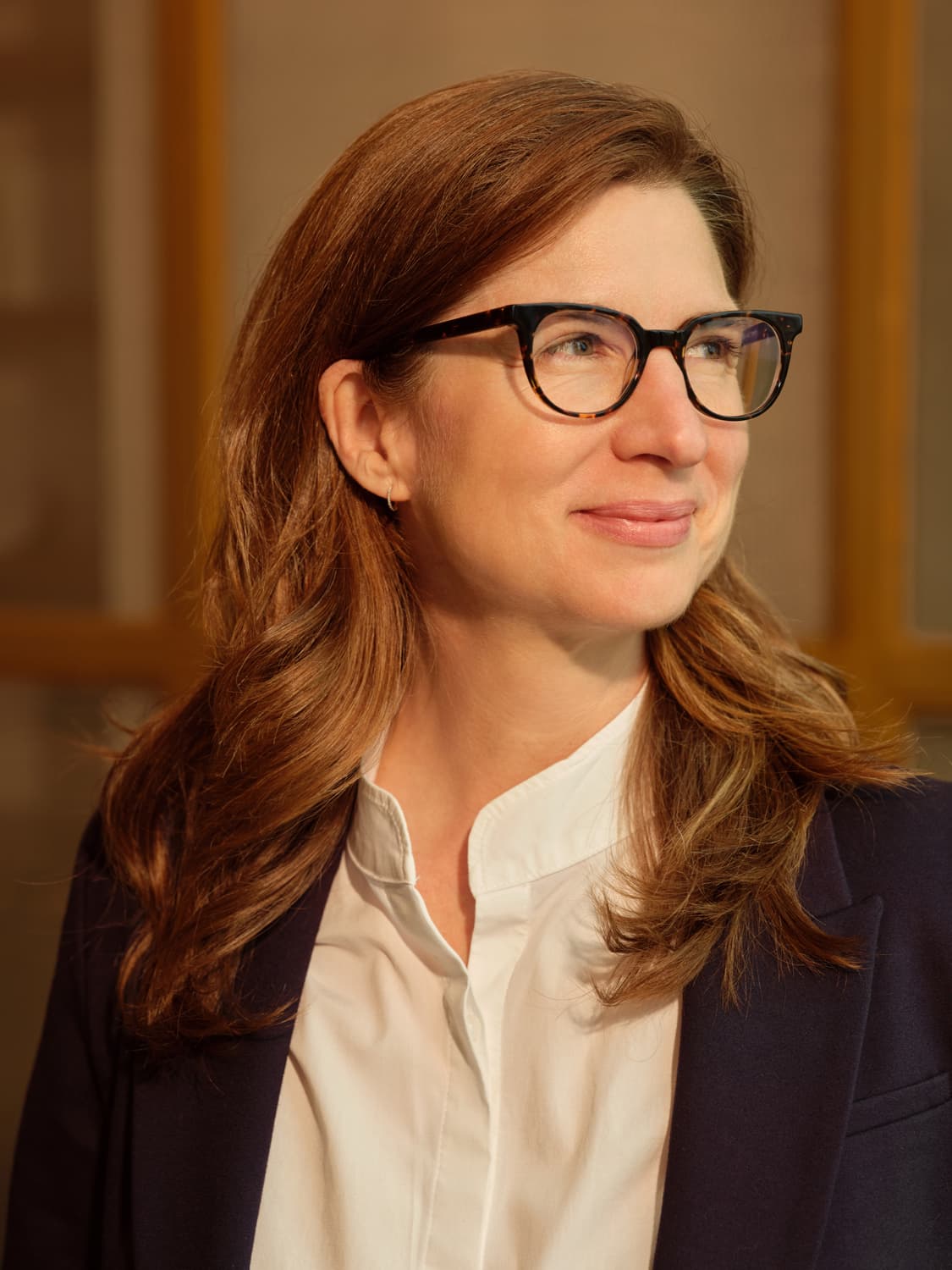 Woman with long auburn hair and tortoiseshell glasses in a navy blazer over a white collared shirt, smiling slightly and looking off camera in warm natural light