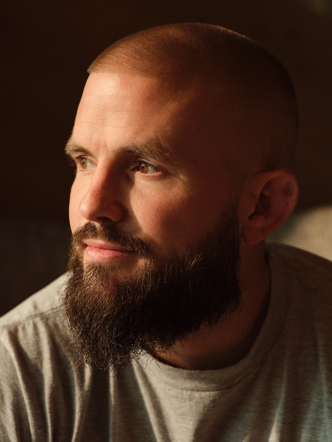 Man with a shaved head and full dark beard in a heathered gray t-shirt, gazing off camera with directional warm light and a dark backdrop behind him