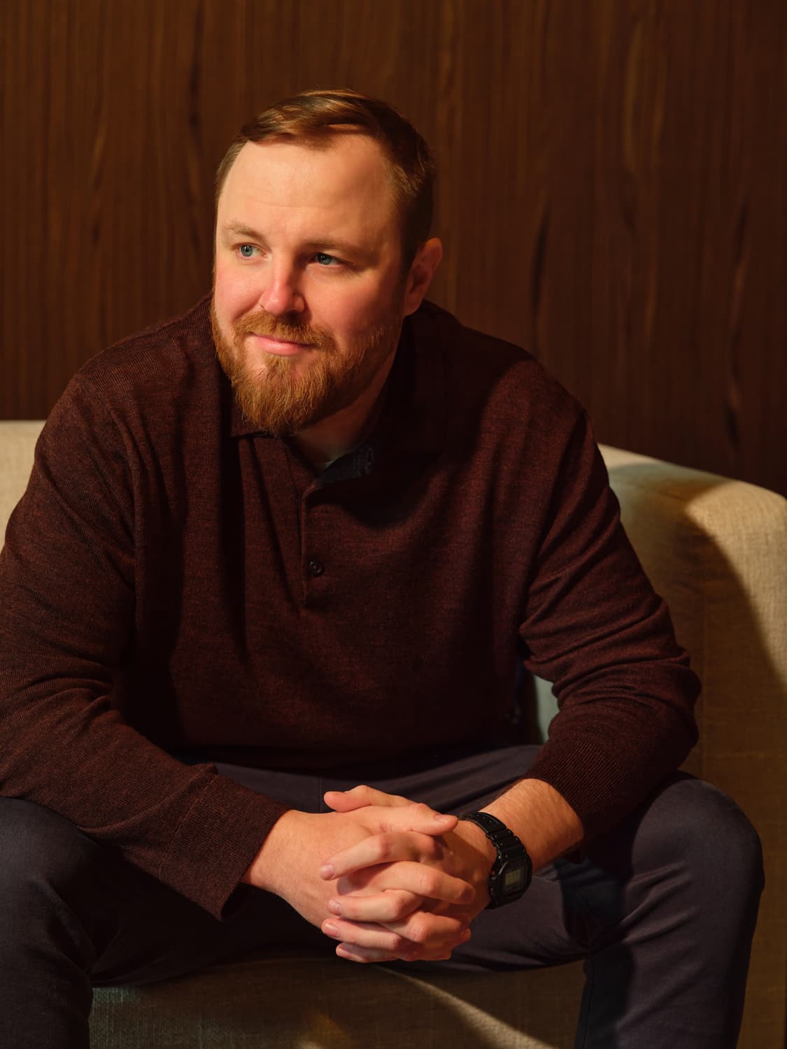 Man with a reddish brown beard in a burgundy wool sweater seated in a chair, hands clasped, looking off camera with warm side light against a dark wood paneled wall