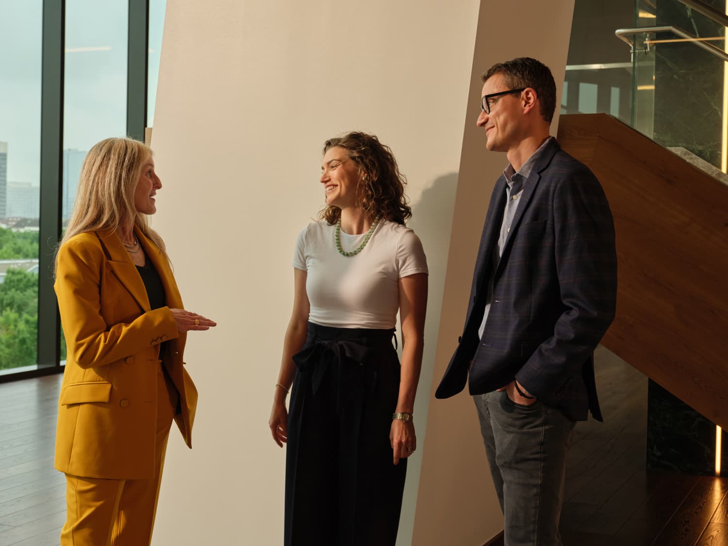 Three people in conversation in a sunlit Houston office lobby, a woman in a yellow suit gesturing while a woman in a white t-shirt with green beaded necklace and a man in a navy plaid blazer listen, downtown skyline visible through floor to ceiling windows