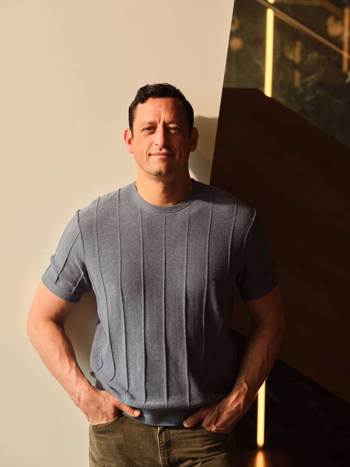 Man with dark hair in a textured gray ribbed t-shirt standing with hands in pockets against a cream wall, warm sidelight casting a soft shadow