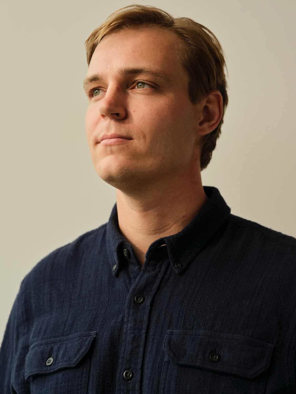 Young man with blonde hair in a navy double pocket workshirt looking up and off camera with soft natural light against a cream toned wall