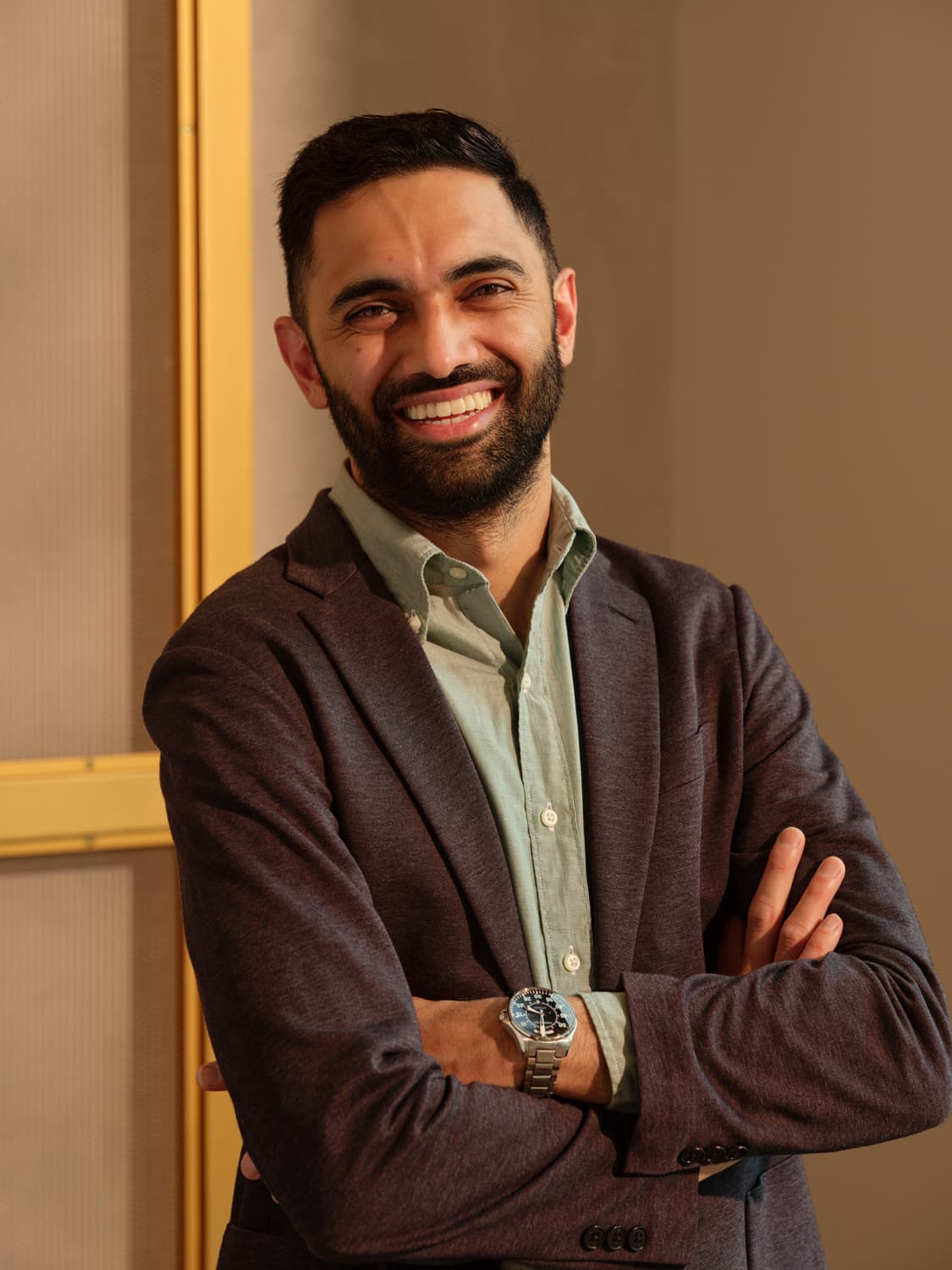 Man with dark hair and beard in a charcoal blazer over a sage green shirt with arms crossed, smiling broadly with warm light against a textured yellow framed backdrop
