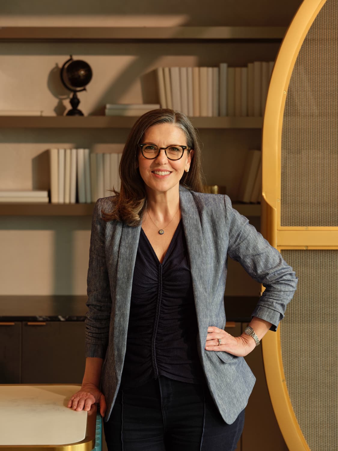 Woman with shoulder length dark hair and tortoiseshell glasses in a chambray blazer over a navy ruched top, standing with hand on hip beside a bookshelf with globe and arched golden screen
