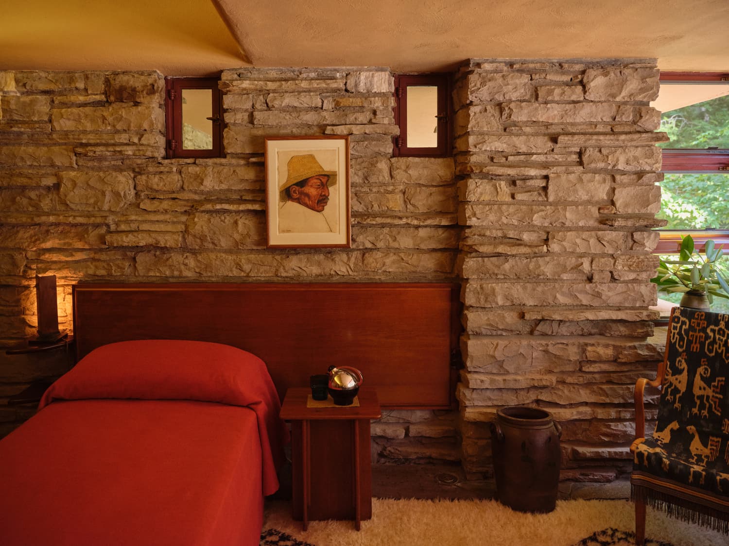 Fallingwater bedroom with red coverlet, stone walls, portrait painting, and small clerestory windows