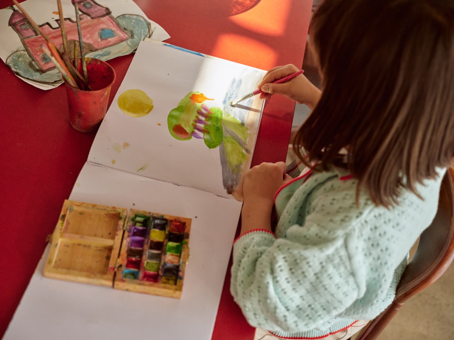 Young girl painting with watercolors at a red table, seen from above