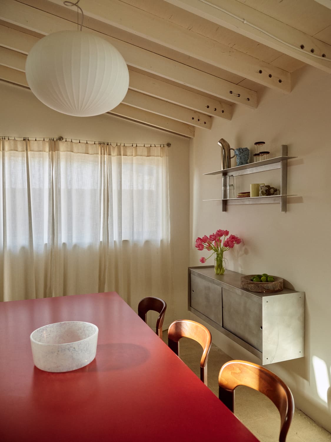 Dining room with red table, wooden chairs, paper lantern, pink tulips, and metal sideboard