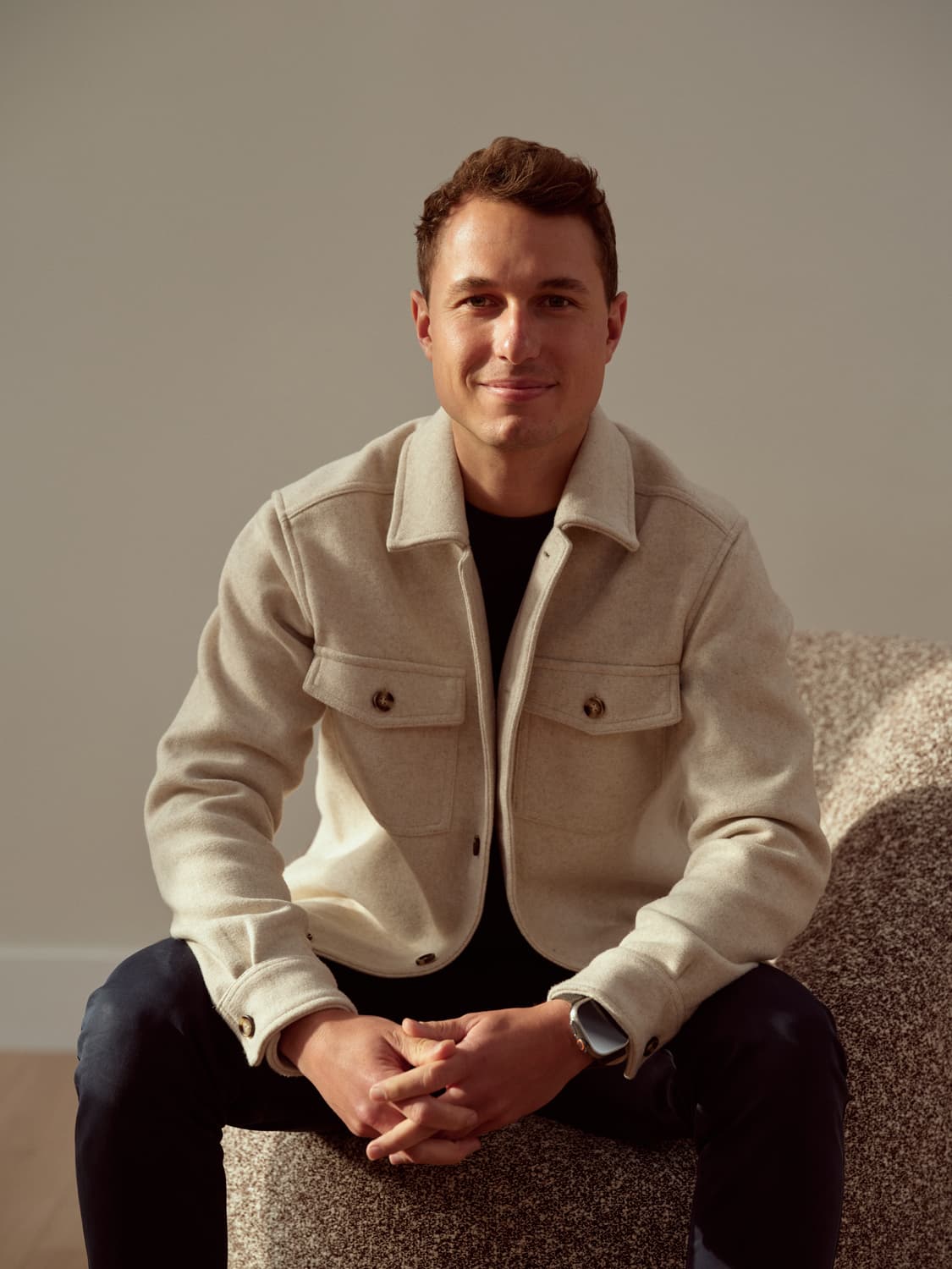 Young man with short brown hair in a cream wool shirt jacket seated on a textured sofa, smiling with hands clasped together