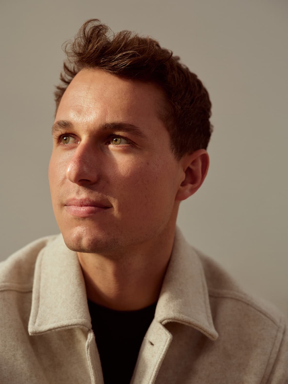 Young man with wavy brown hair in a cream wool jacket over a black tee, looking up with soft studio light on his face