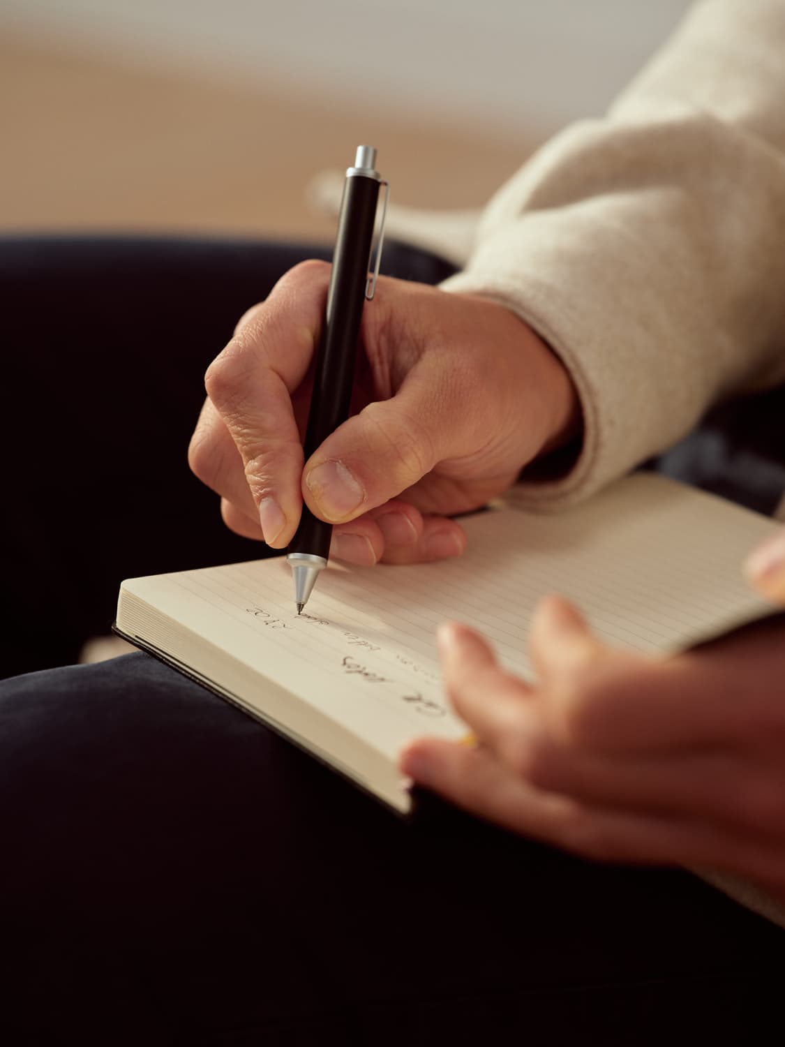 Close-up of hands writing in a lined notebook with a black pen, cream sweater sleeve visible in warm ambient light