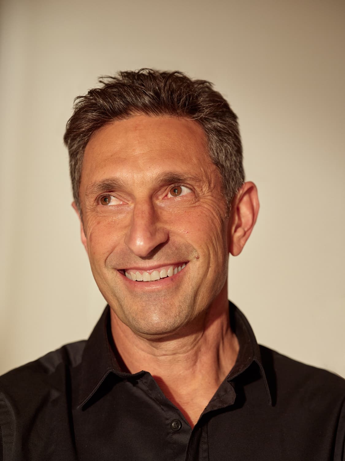 Man with gray-flecked dark hair in a black shirt smiling broadly and looking off camera against a light backdrop