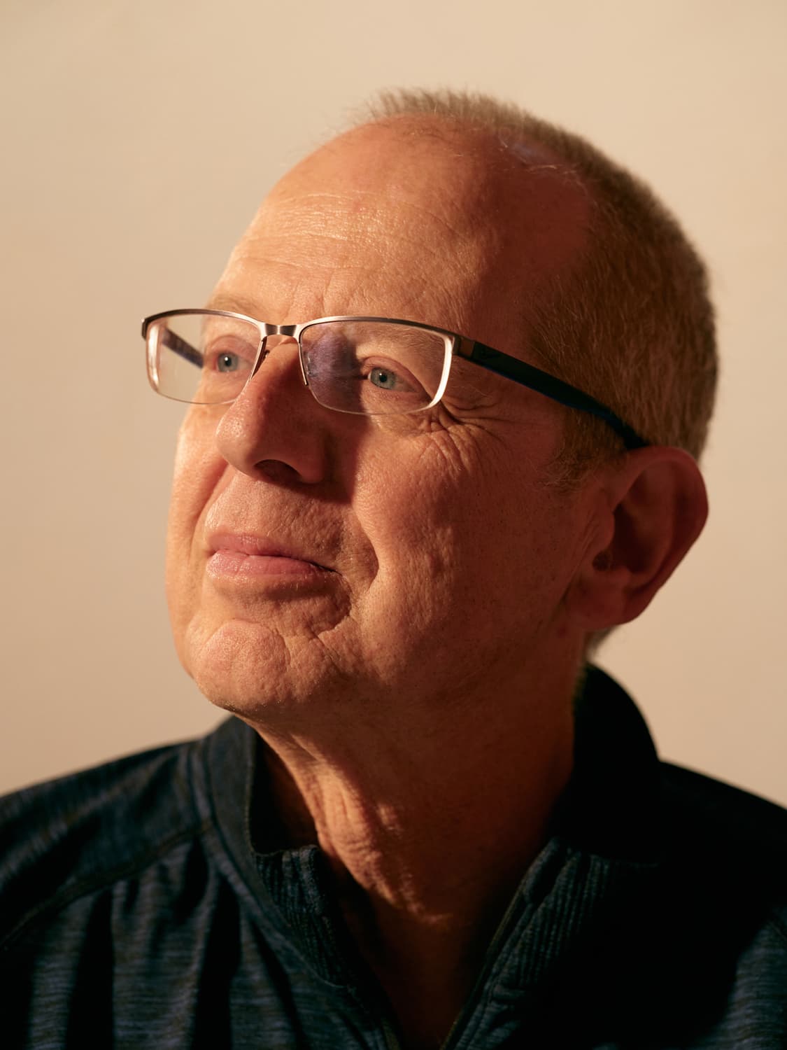 Older man with short hair and rectangular glasses in a heathered blue shirt, warm light on his face as he gazes upward