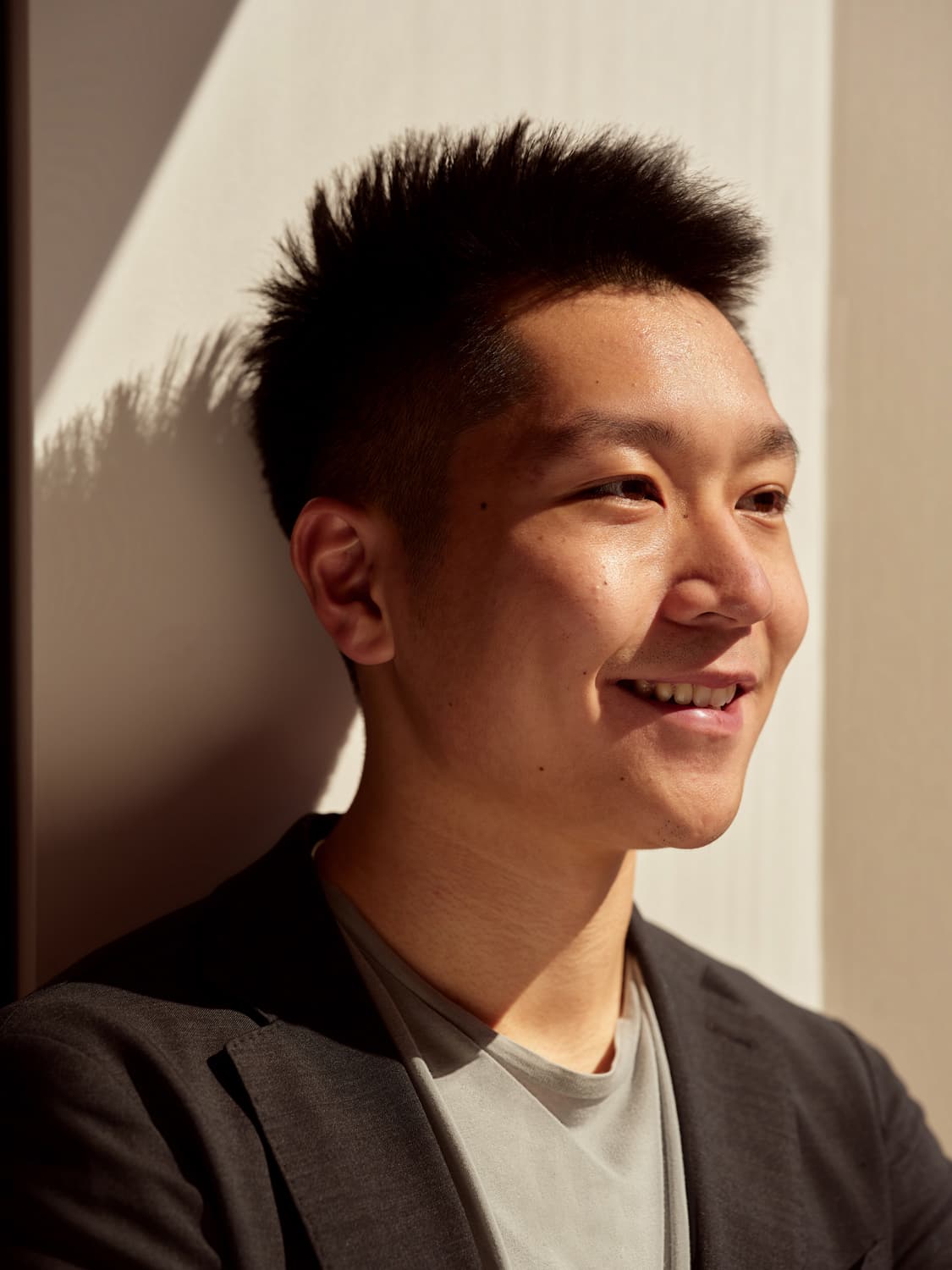 Young man with spiky dark hair in a gray blazer and tee smiling in profile, warm sunlight casting shadows on the wall behind