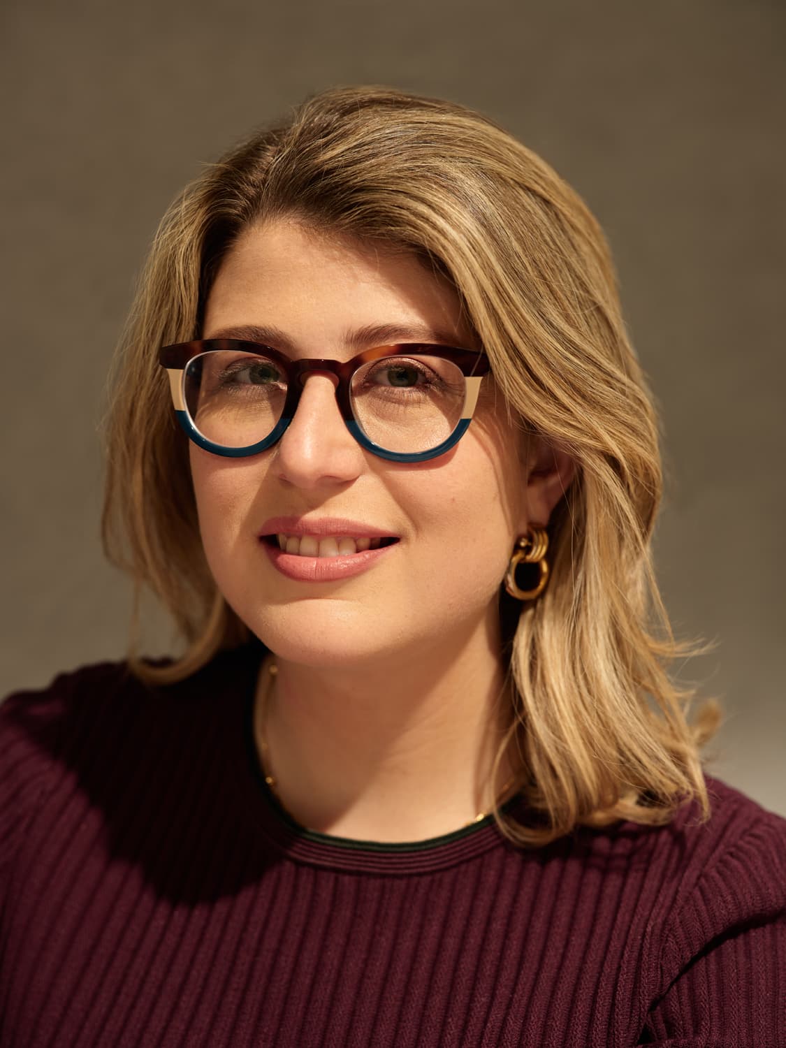Woman with blonde wavy hair wearing colorful two-tone glasses, gold hoop earrings, and a burgundy ribbed sweater