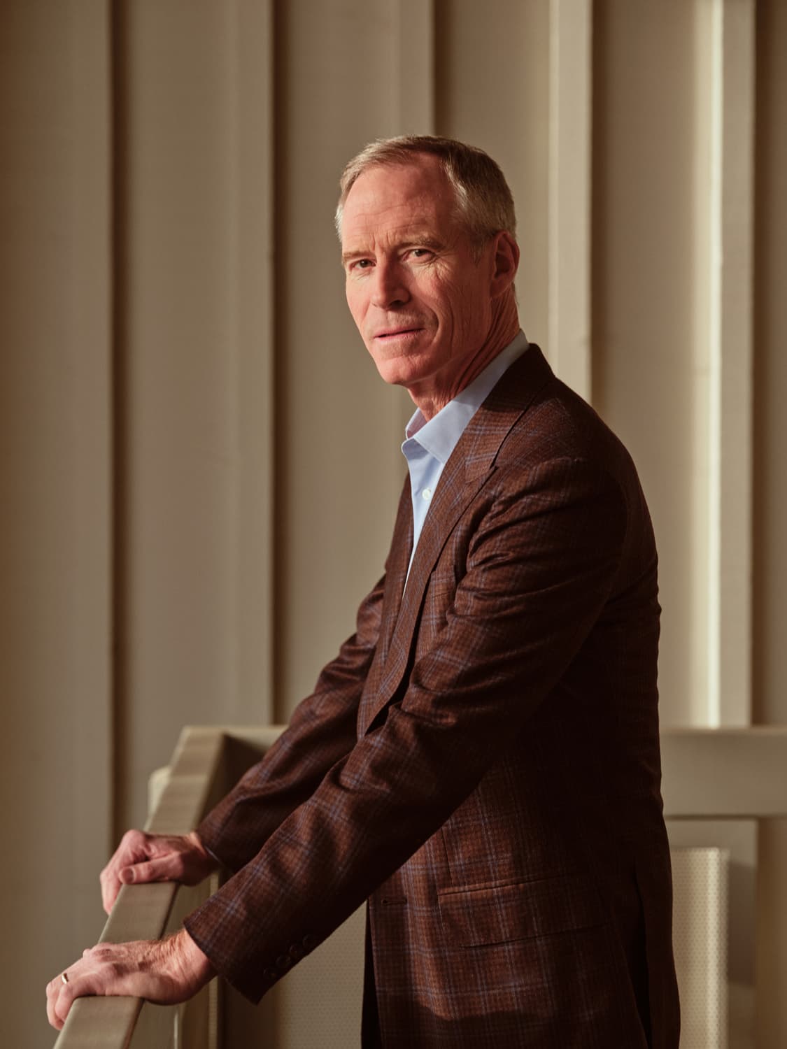 Man with light hair in a brown plaid sport coat and blue shirt leaning on a chair, dramatic window light in an office lobby