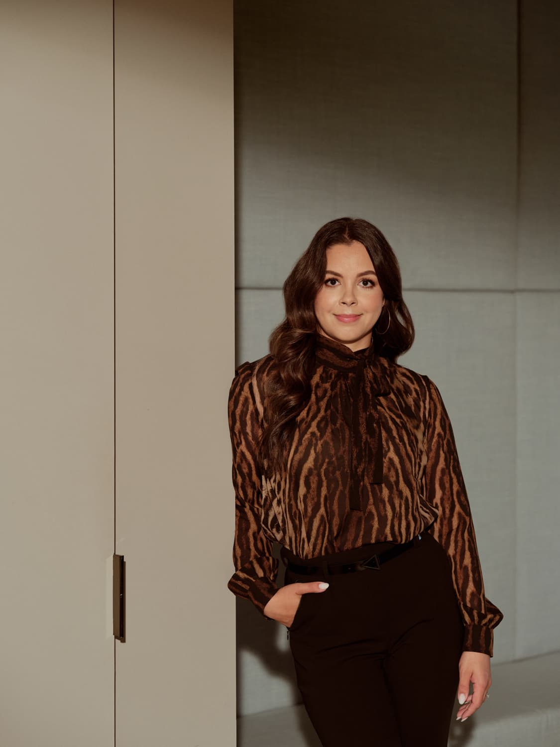 Woman with dark wavy hair in a leopard print bow blouse and black trousers, standing beside a closet door in a modern office
