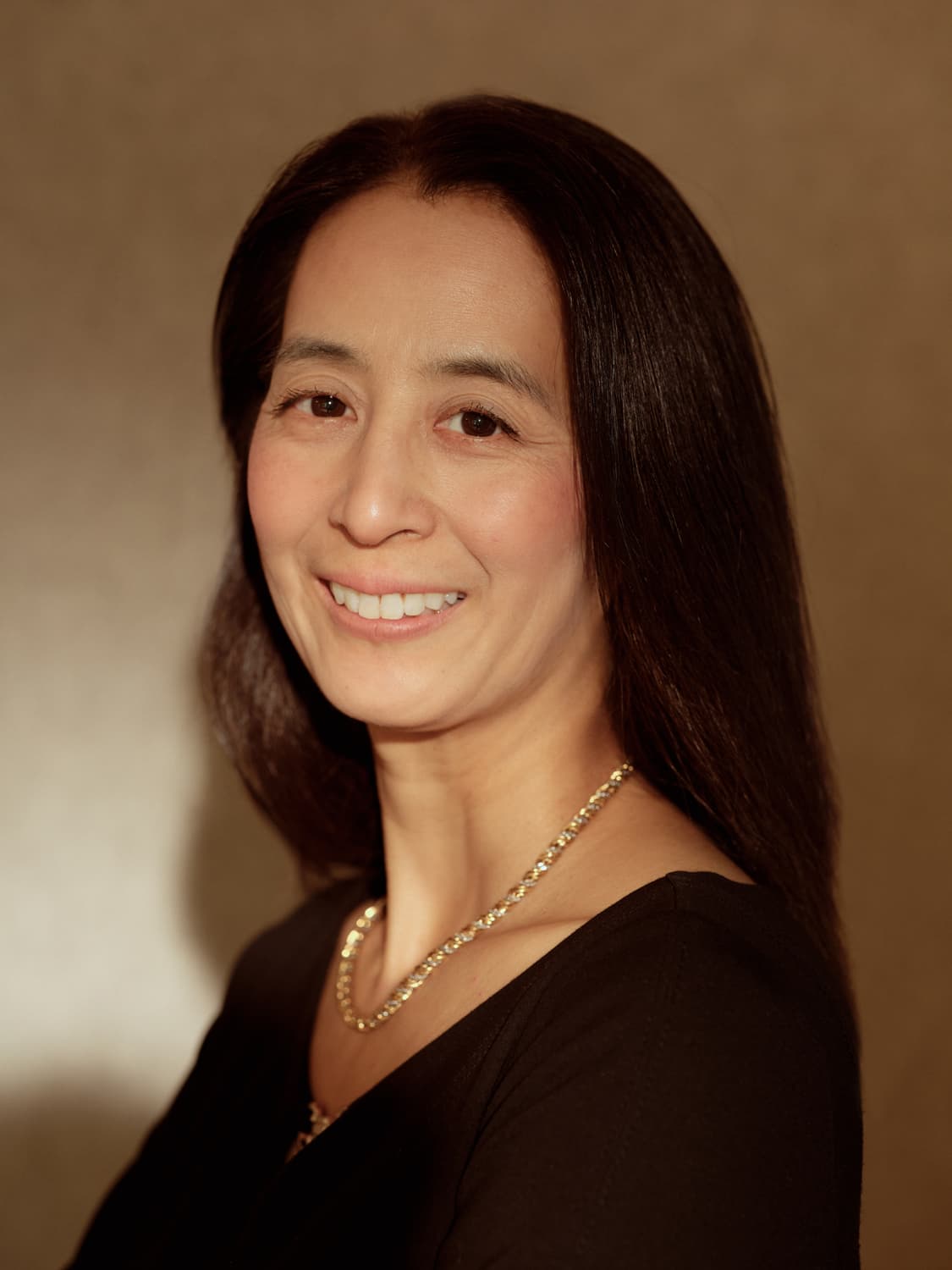Woman with long dark hair in a black v-neck top and gold chain necklace smiling warmly against a brown toned backdrop