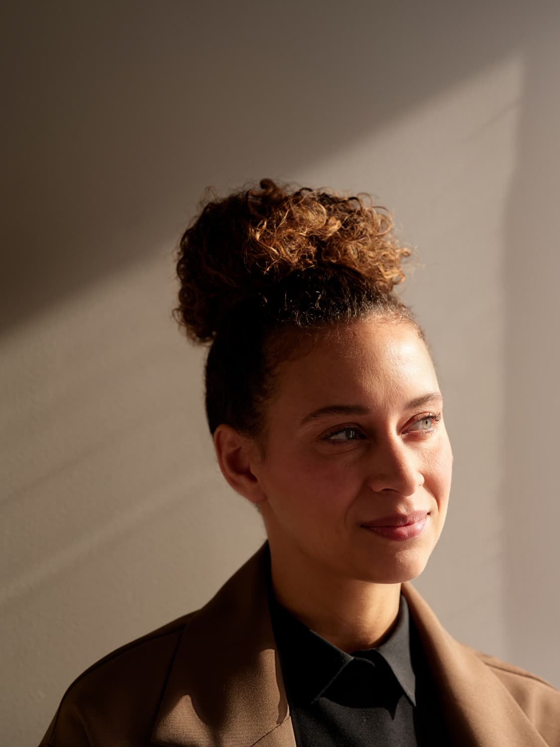 Woman with curly hair in a bun wearing a brown blazer and black shirt, smiling and looking off camera with natural window light