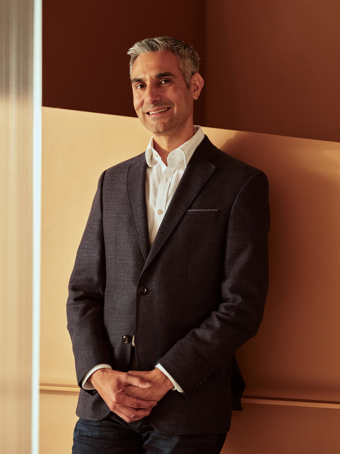 Man with silver hair in a charcoal blazer and white dress shirt leaning against a warm terracotta wall, smiling off camera