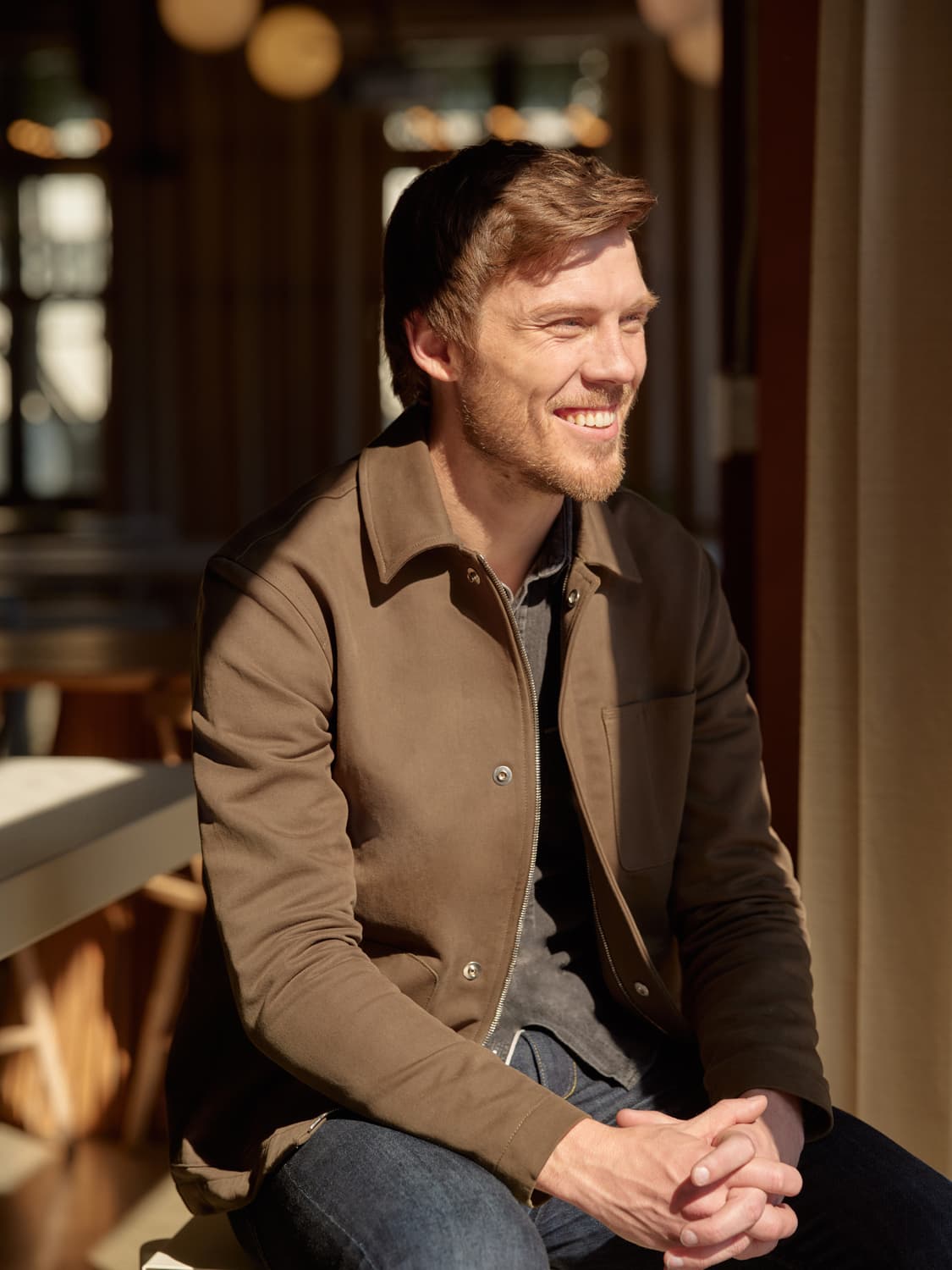 Man with reddish beard in a brown jacket and jeans seated in a cafe-like space, laughing with warm backlight behind him