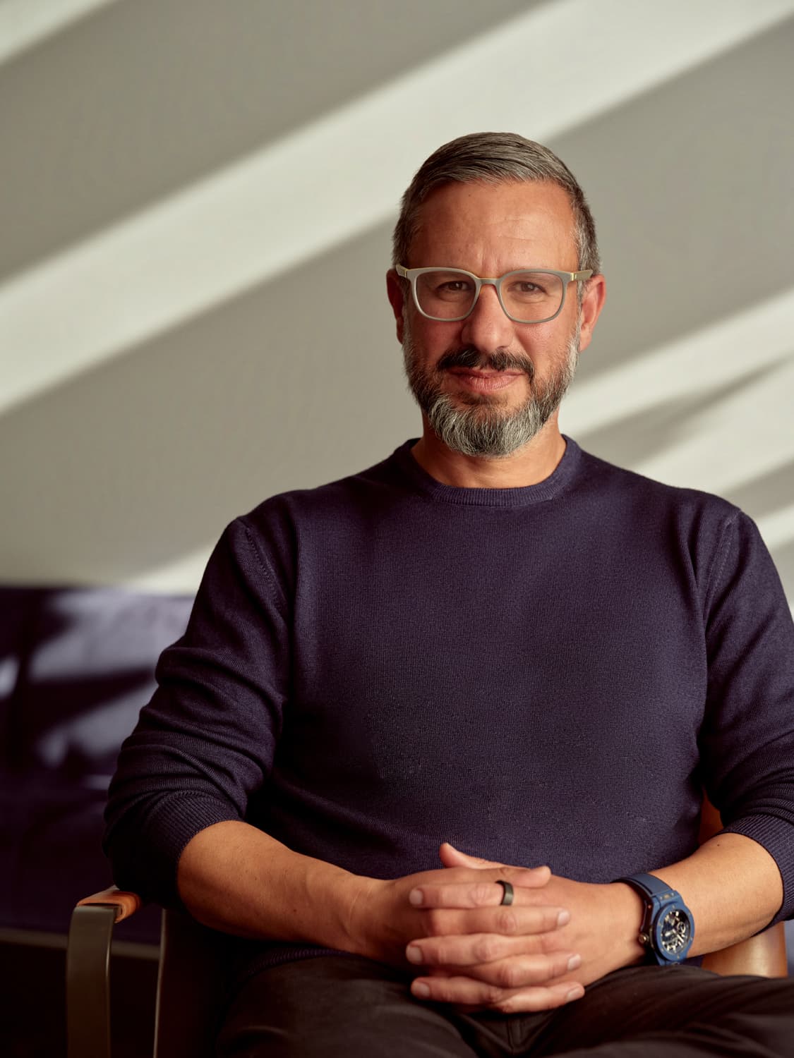 Man with glasses and a salt-and-pepper beard in a navy crewneck sweater seated with hands clasped, diagonal light behind him