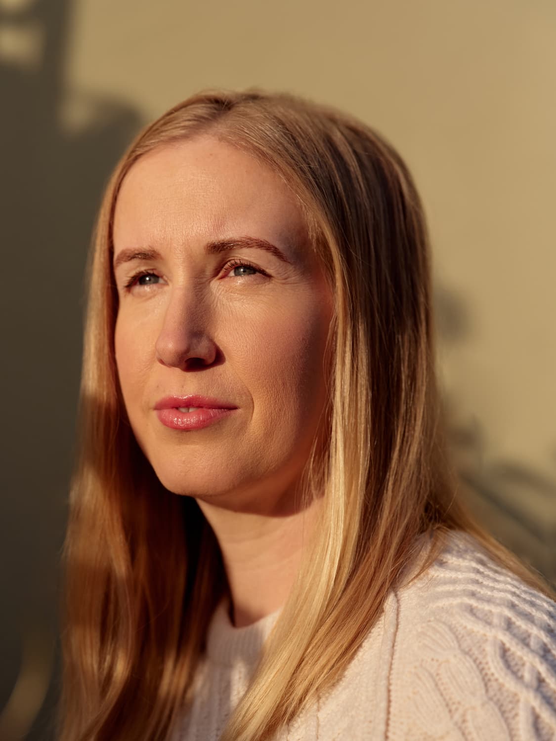 Woman with long straight blonde hair in a cream cable-knit sweater, soft golden light across her face as she looks off camera