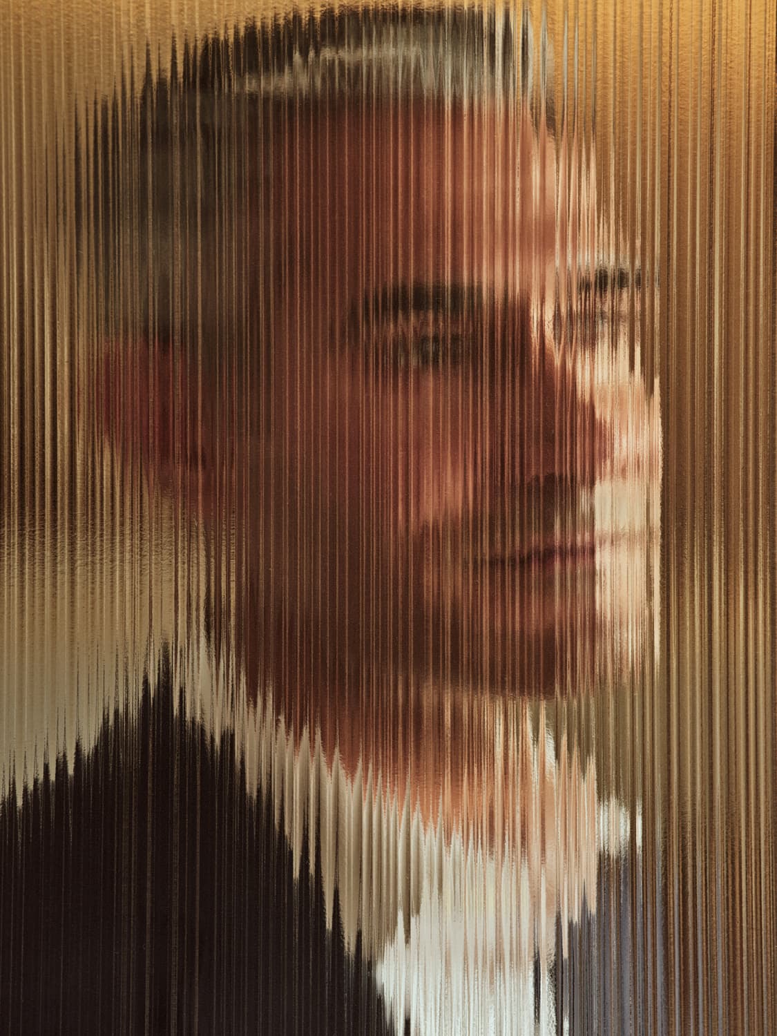 Abstract portrait of a person seen through textured reeded glass, creating vertical streaks of warm golden tones