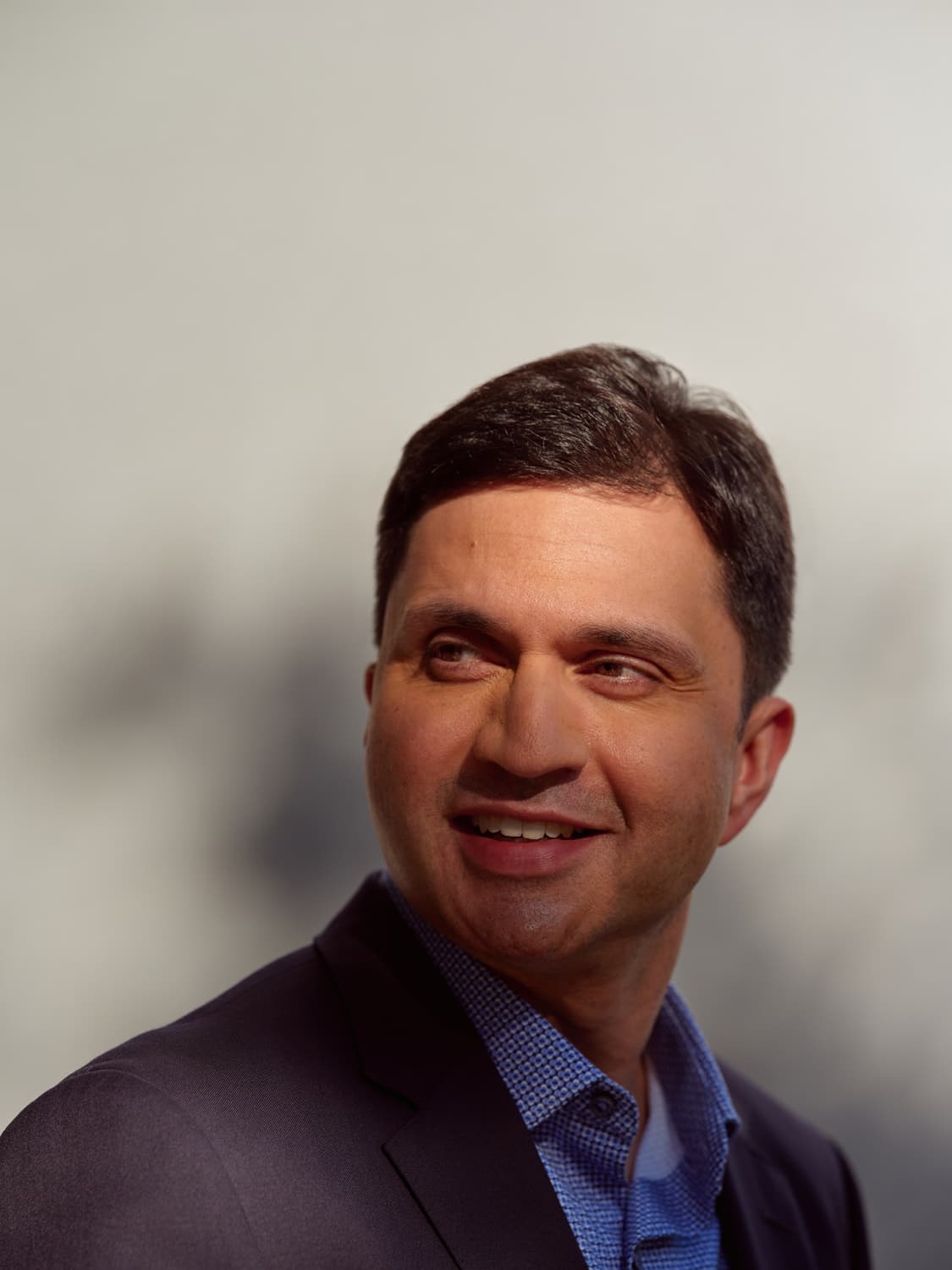 Man in a navy blazer and blue patterned shirt smiling and looking off camera with a soft blurred background