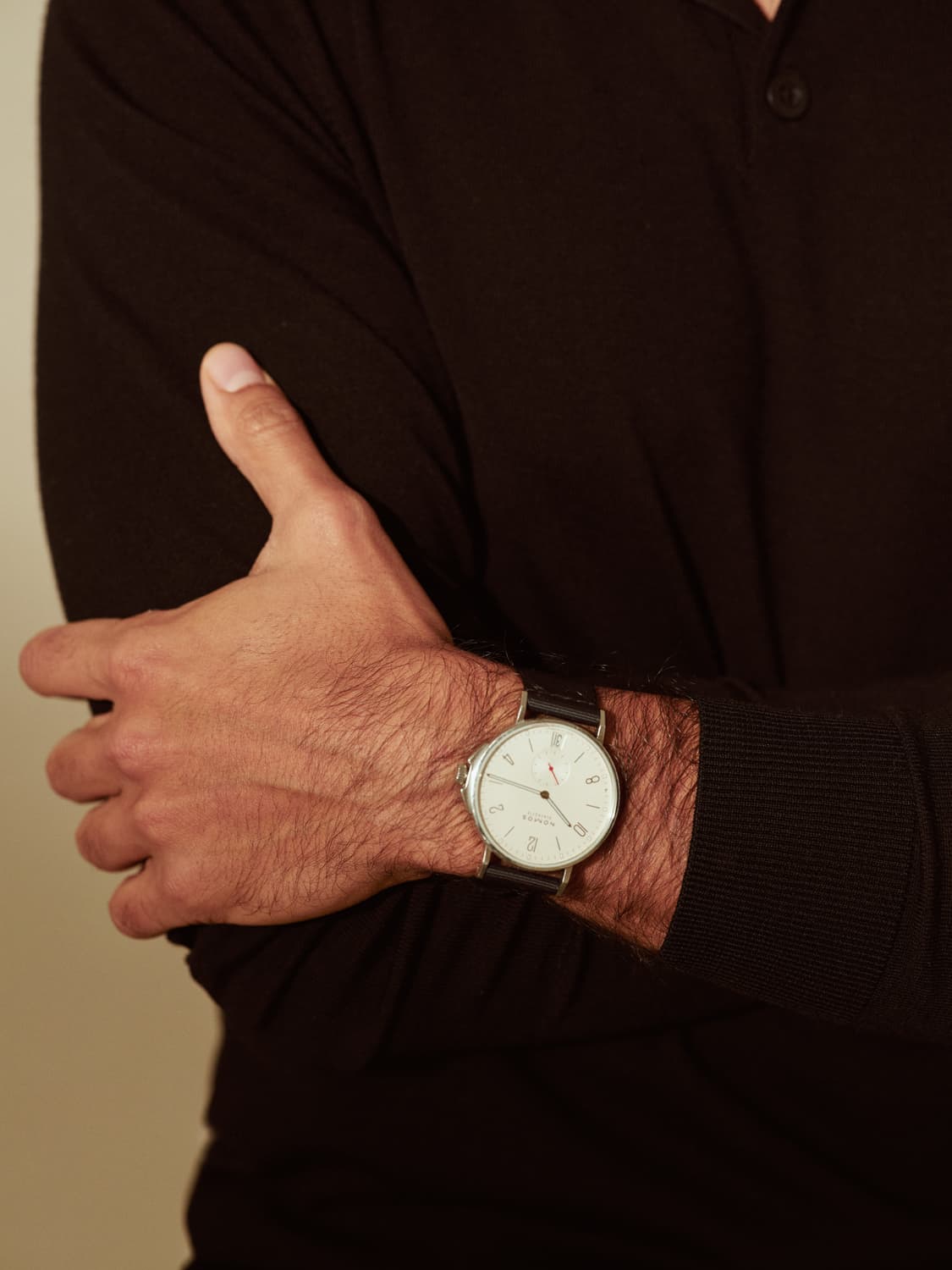 Close-up detail of crossed arms in a dark knit polo showing a minimalist white-dial wristwatch with a leather strap