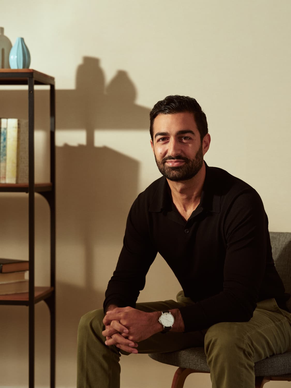 Man with dark beard in a black polo seated on a gray couch, hands clasped, with a bookshelf behind him in warm office light