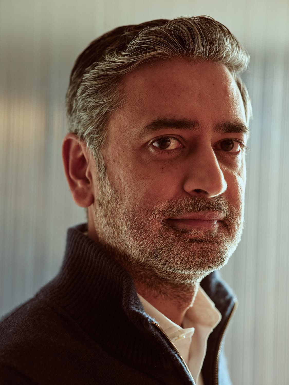 Close-up portrait of a bearded man with gray-streaked hair in a dark quarter-zip and cream collar, looking into the camera