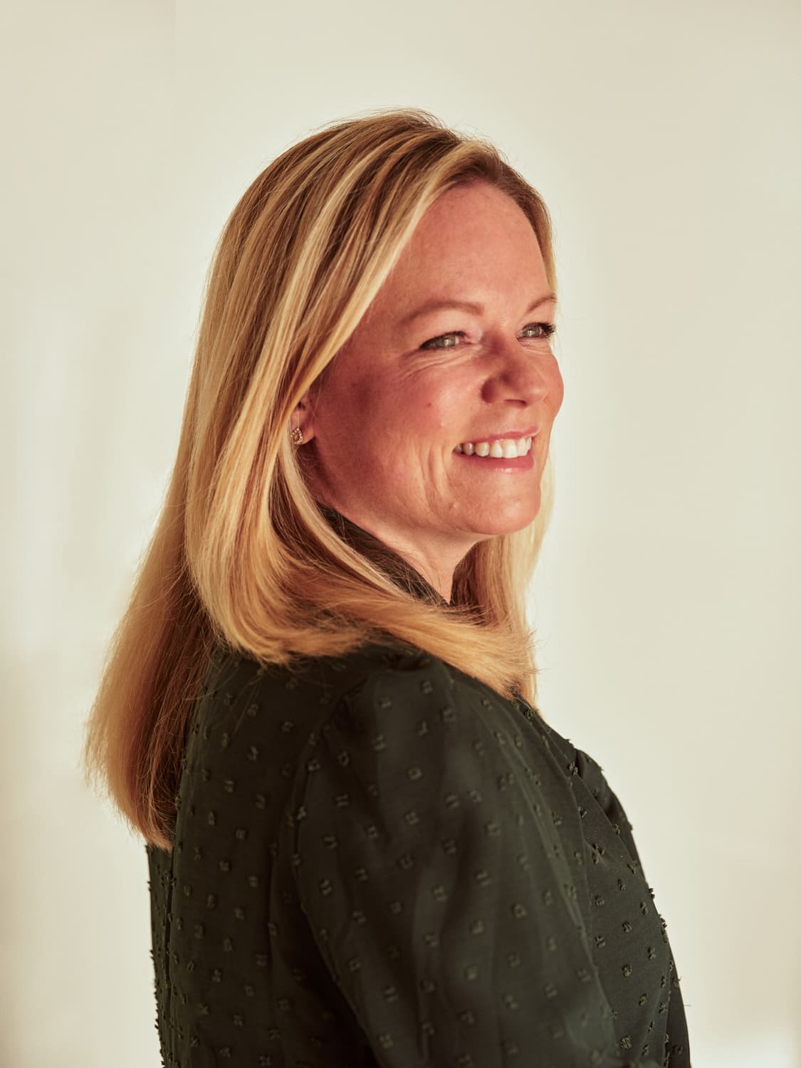 Woman with straight blonde hair looking over her shoulder and smiling in a dark textured blouse against a bright white background