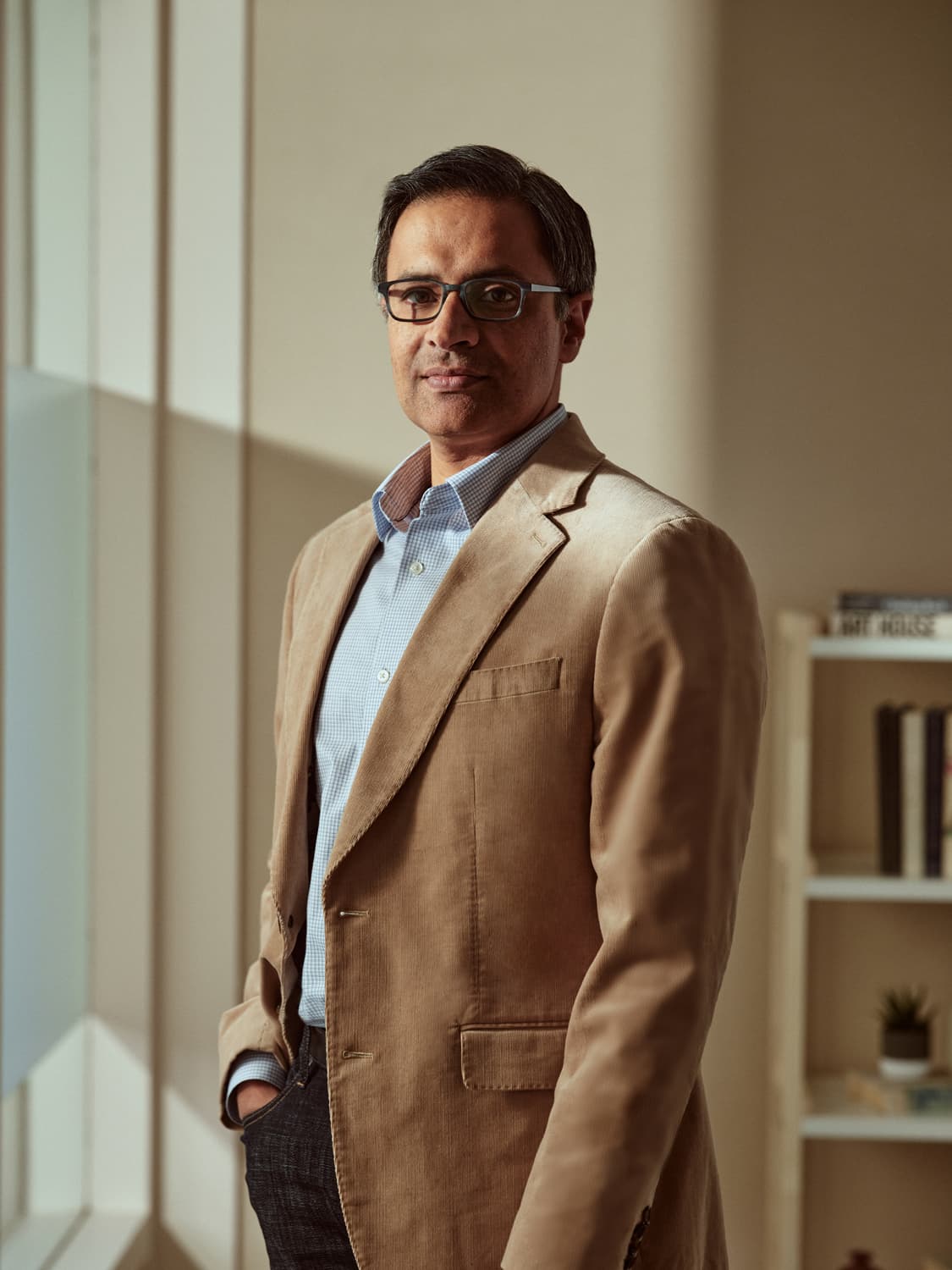 Man in a tan corduroy blazer and light blue check shirt standing by a window with a bookshelf, hand in pocket