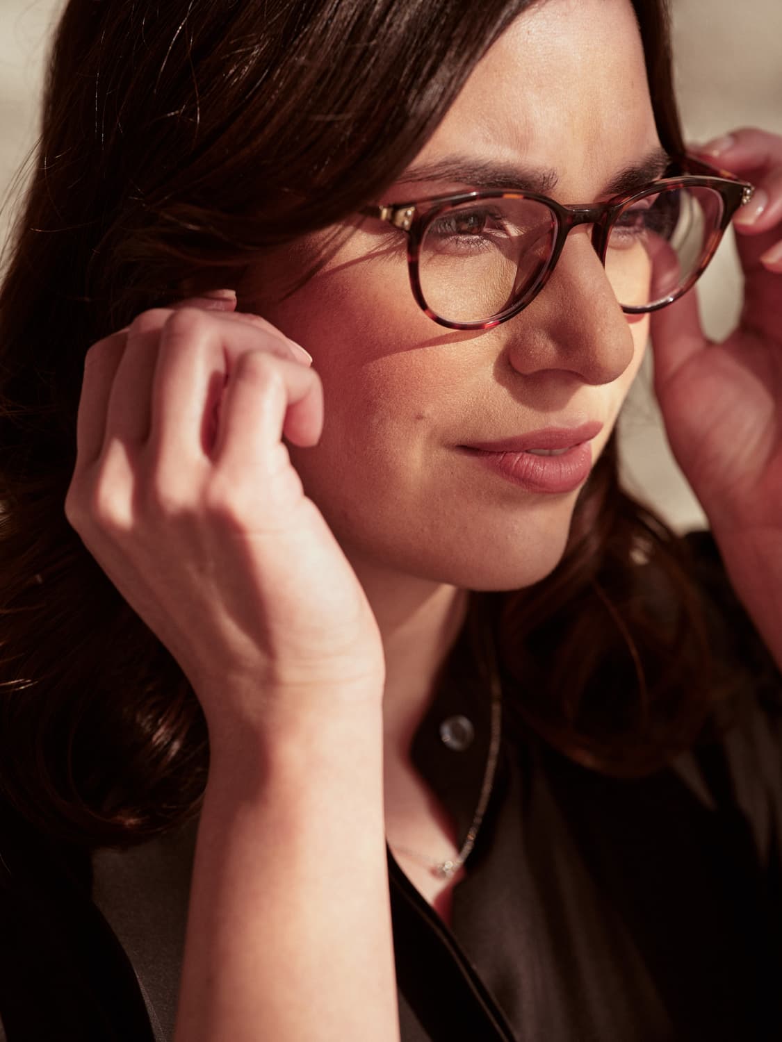 Close-up of a woman with dark hair and tortoiseshell glasses adjusting her frames, warm light falling across her face