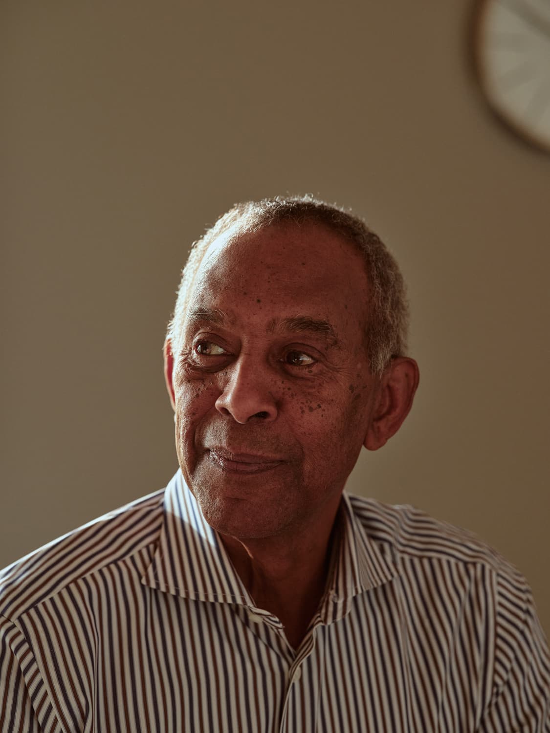 Older man in a striped button-down shirt seated in a conference room with a wall clock, warm light on his face