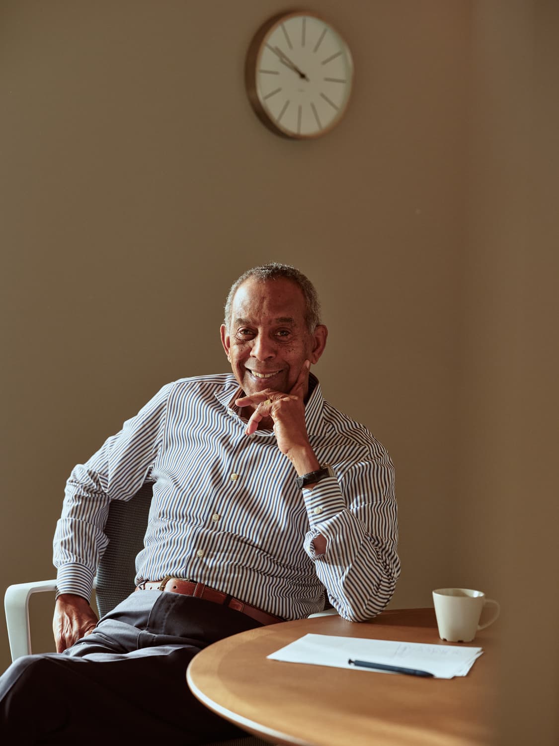 Older man in a striped shirt smiling with hand to his chin, seated at a round table with a coffee cup and wall clock above