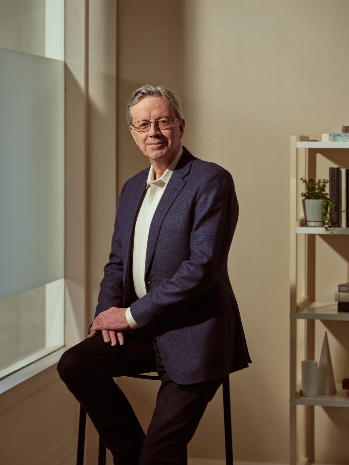 Older man with glasses in a navy blazer and cream open-collar shirt perched on a stool beside a bookshelf with plants