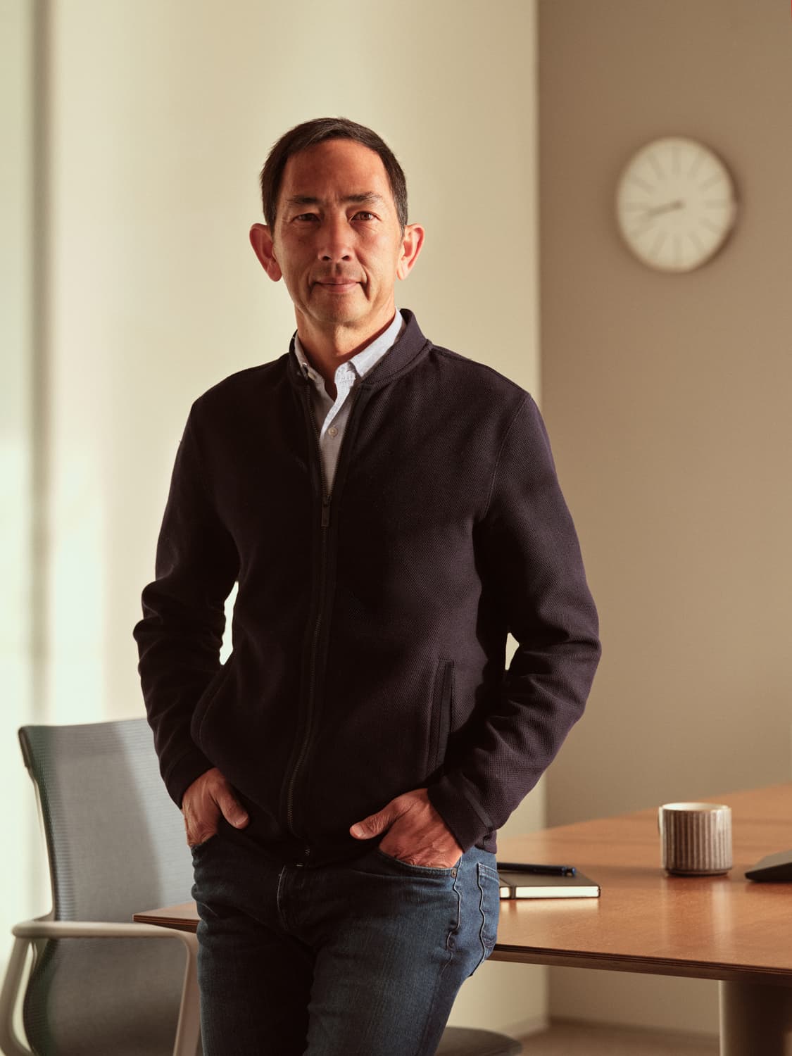 Man in a dark zip-up jacket and light button-down standing with hands in pockets beside a conference table with a wall clock
