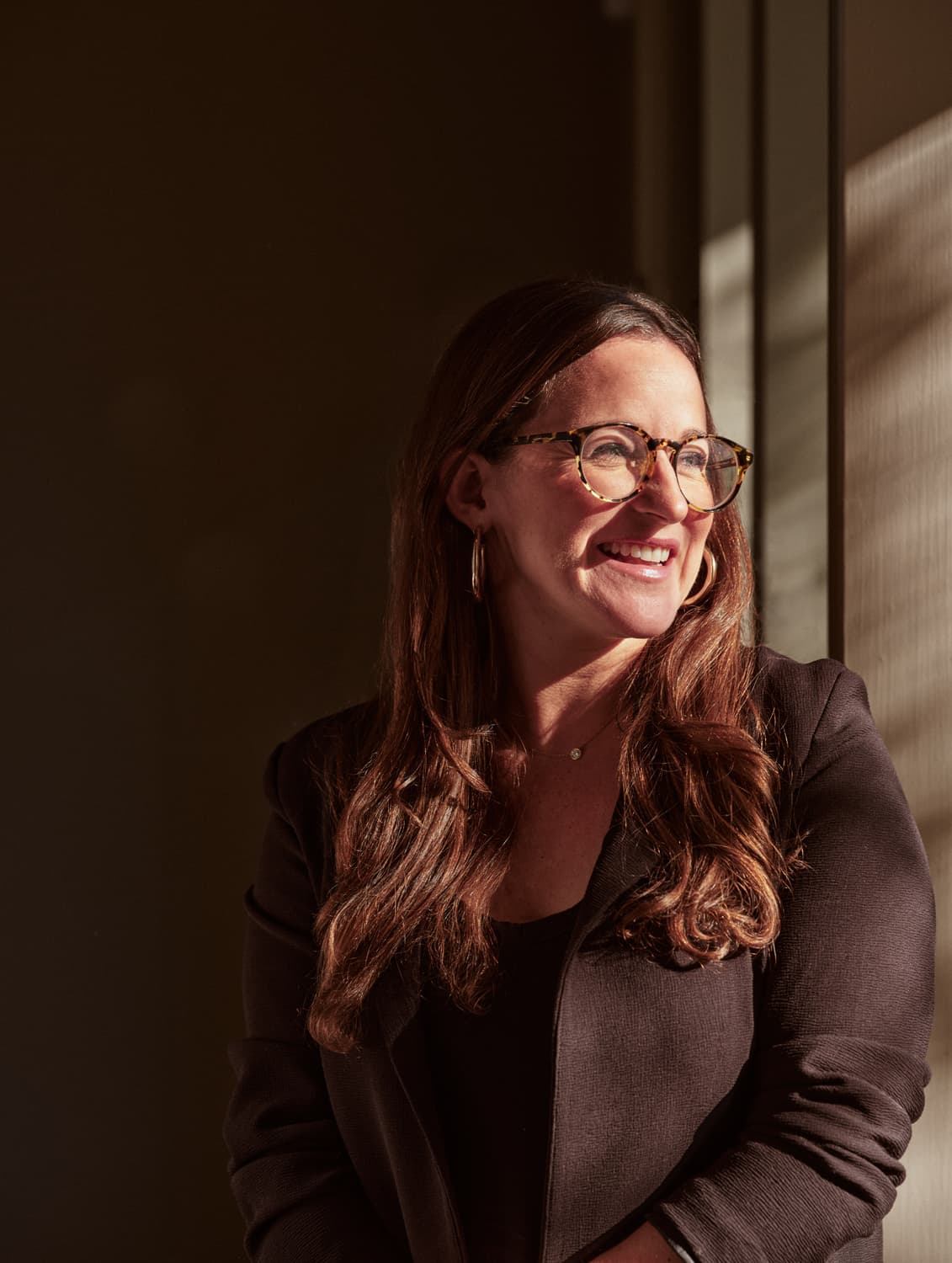 Woman with long brown hair and tortoiseshell glasses laughing in golden window light, wearing a dark blazer over a black top