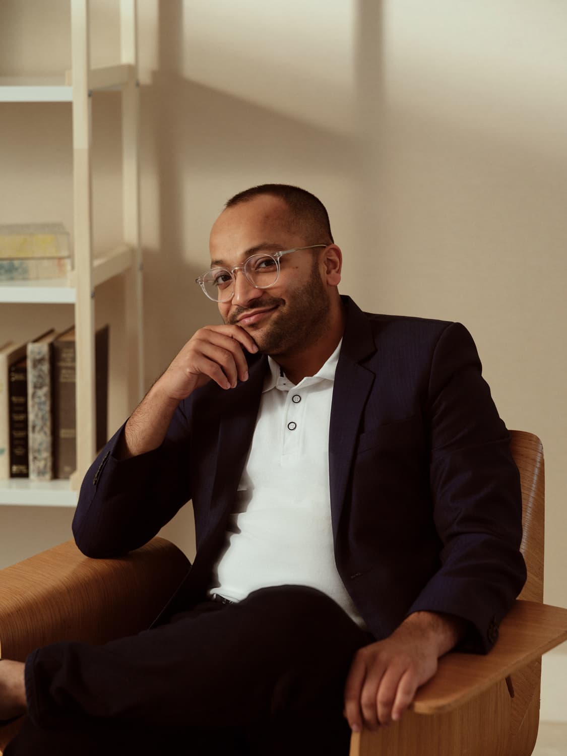 Man with clear-framed glasses in a navy blazer and white polo seated in a wooden chair, hand on chin, with a bookshelf behind