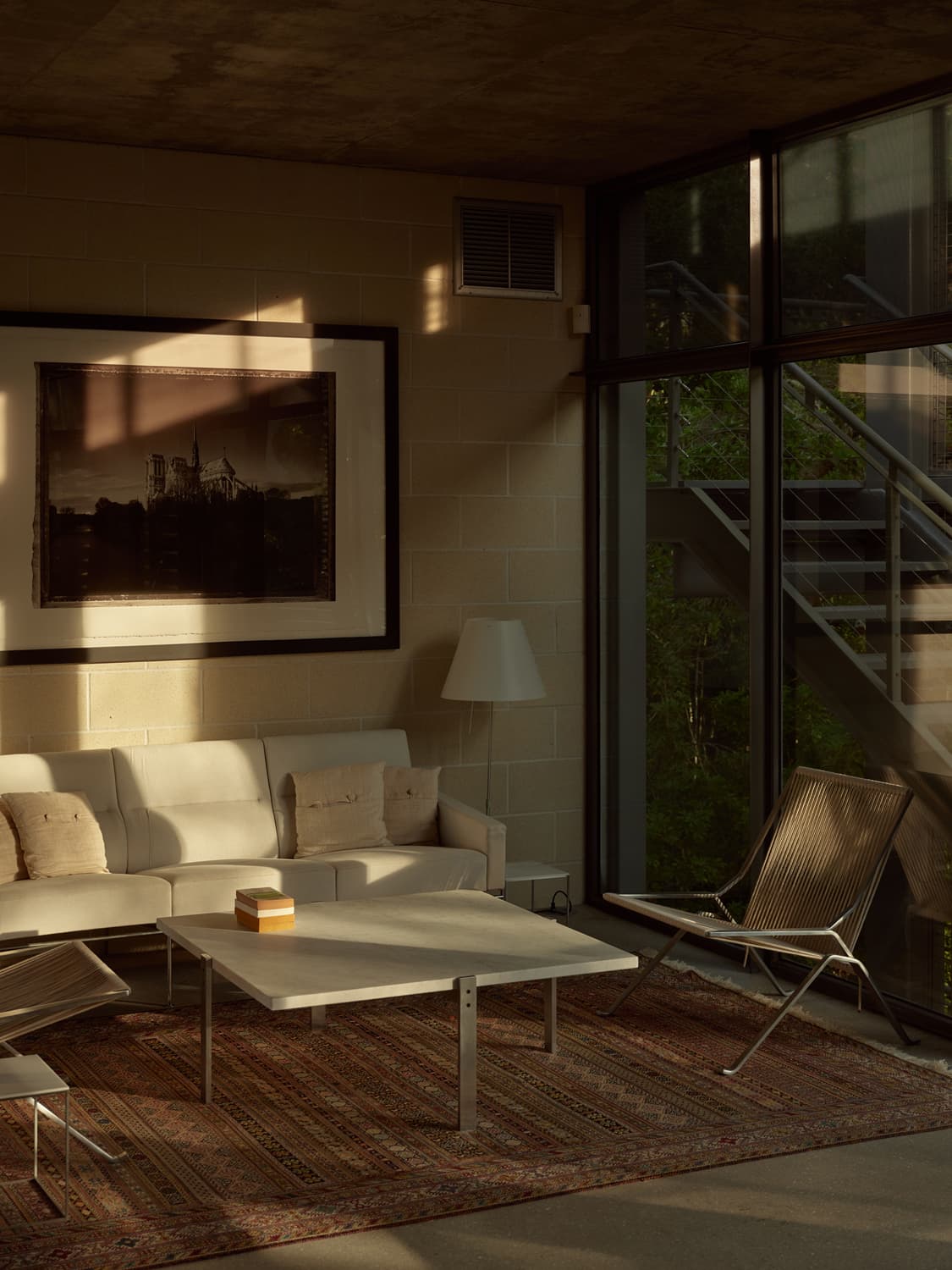 Living room corner with white sofa, large framed photograph, string chair, and Persian rug in warm light