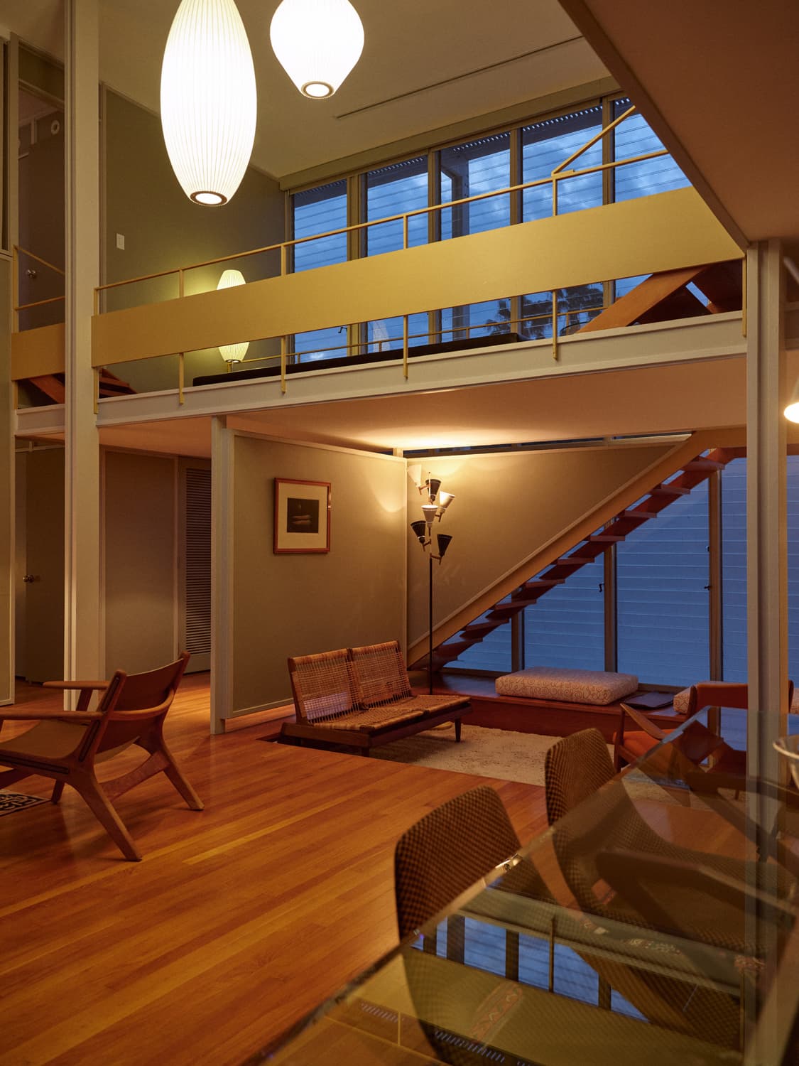 Umbrella House living room at dusk with Nelson pendants, mezzanine, open stair, and warm interior glow