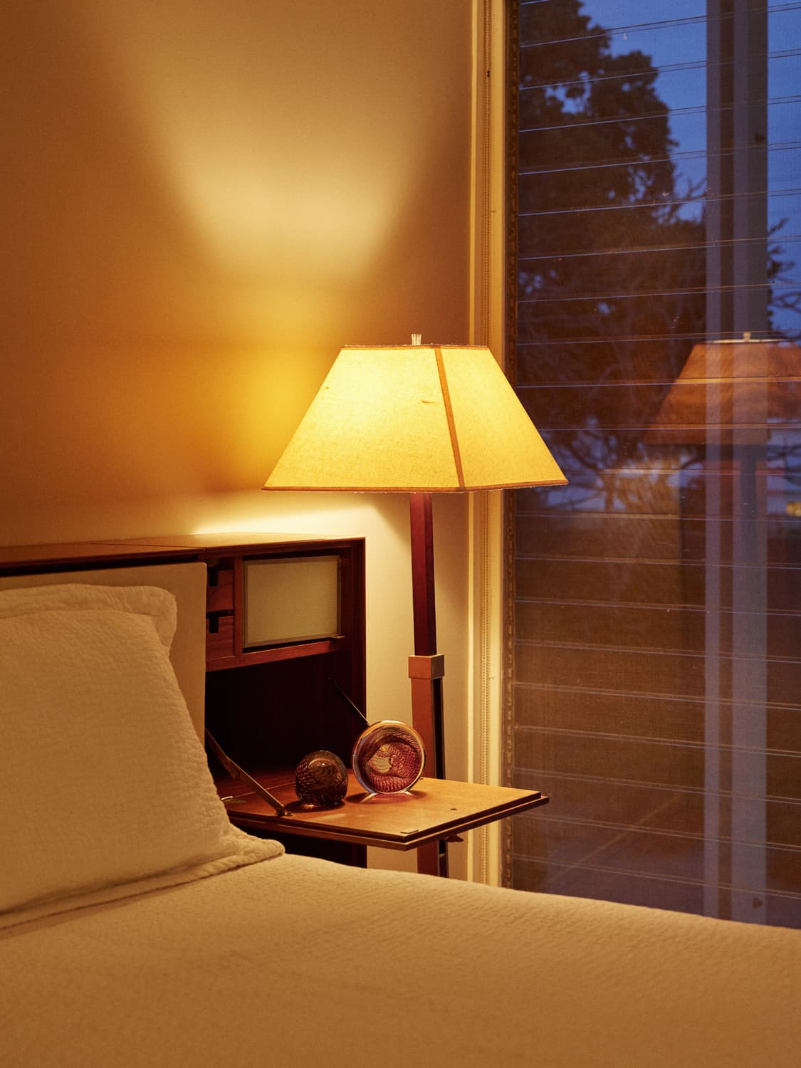 Bedside table lamp glowing warmly next to glass paperweights in the Umbrella House bedroom at twilight