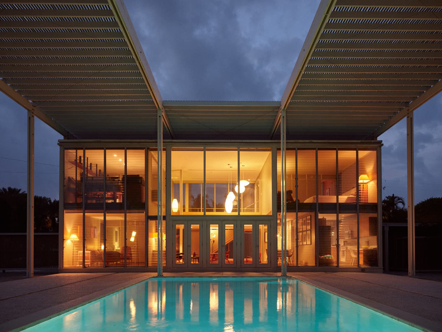 Umbrella House at twilight from the pool with illuminated glass walls and slatted roof silhouette