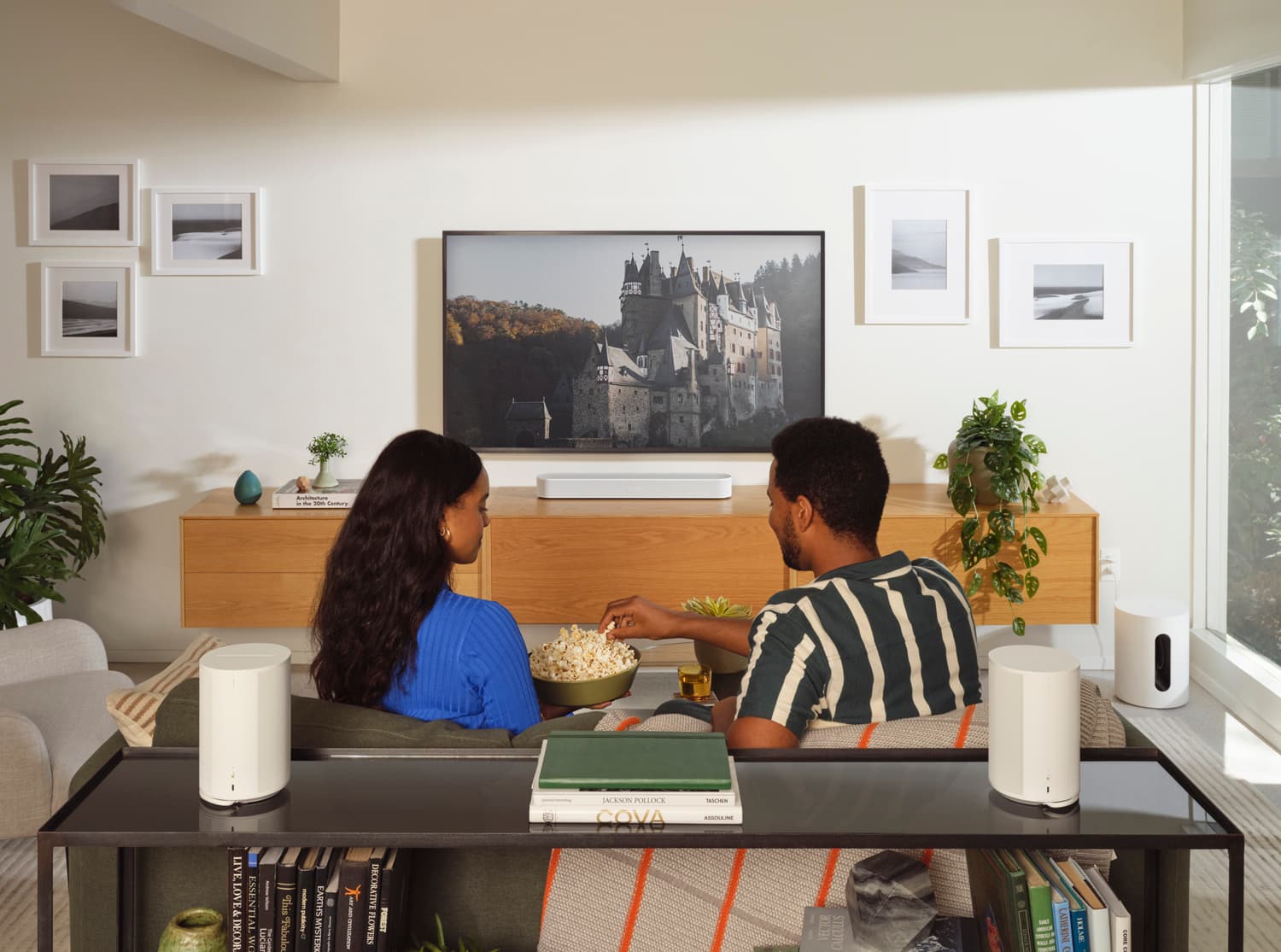 Couple sharing popcorn on a sofa watching TV with two white Sonos Era speakers and a soundbar