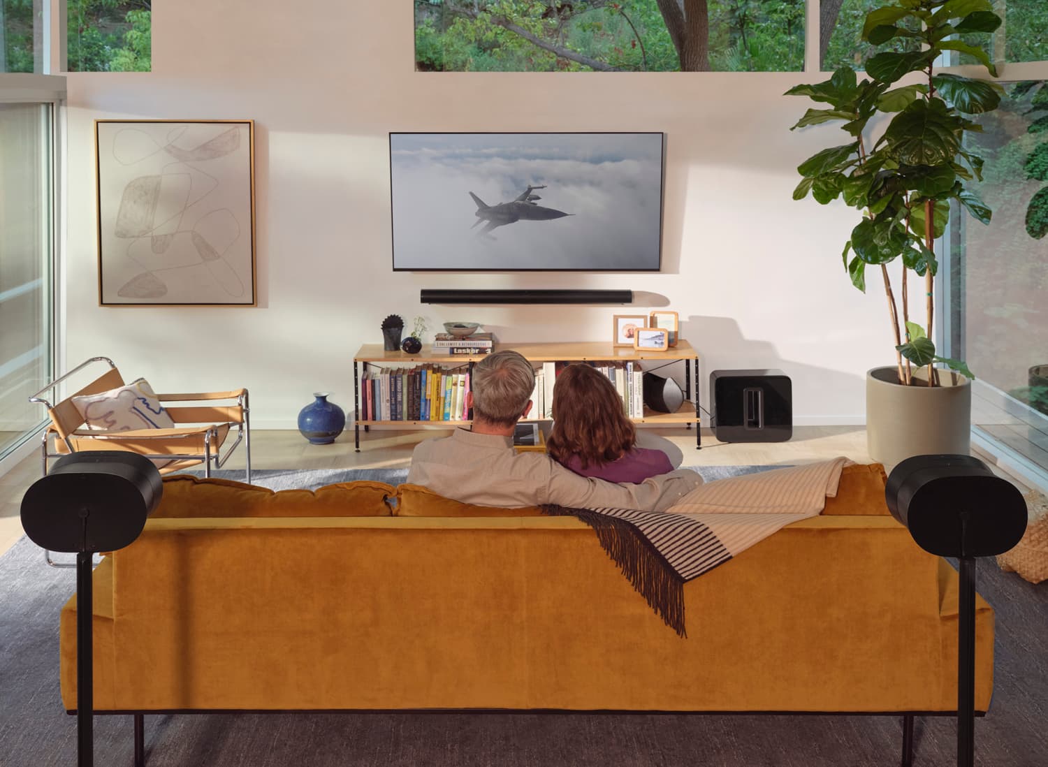 Couple on a mustard velvet sofa watching TV with Sonos Era speakers on stands, soundbar, and sub