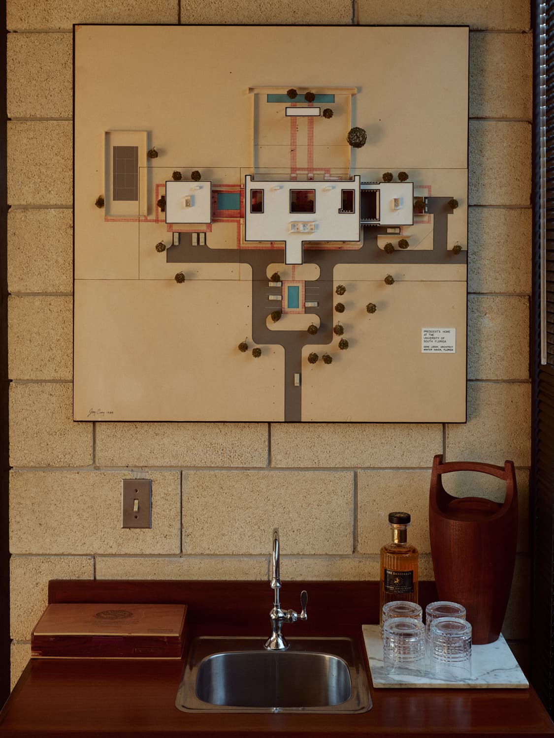 Architectural site model mounted on concrete block wall above a bar sink with whiskey and glasses